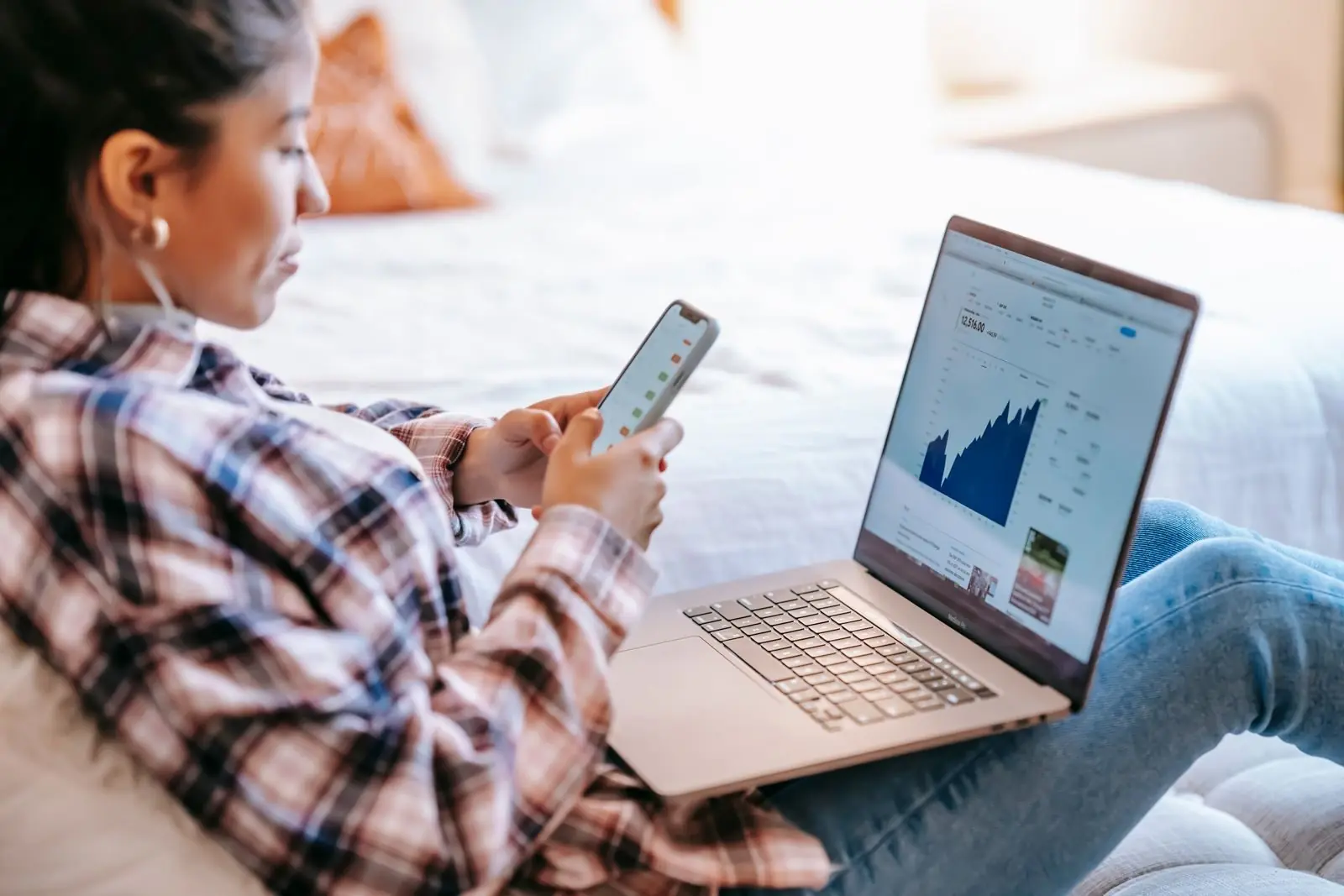 Wanita memeriksa aplikasi trading di ponselnya dengan grafik terbuka di laptopnya